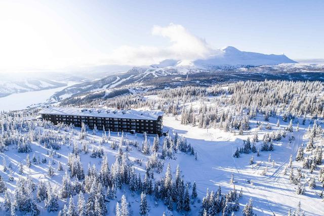 Copperhill Mountain Lodge among snow-laden spruce above Åre, with the ski slopes and the frozen lake beyond